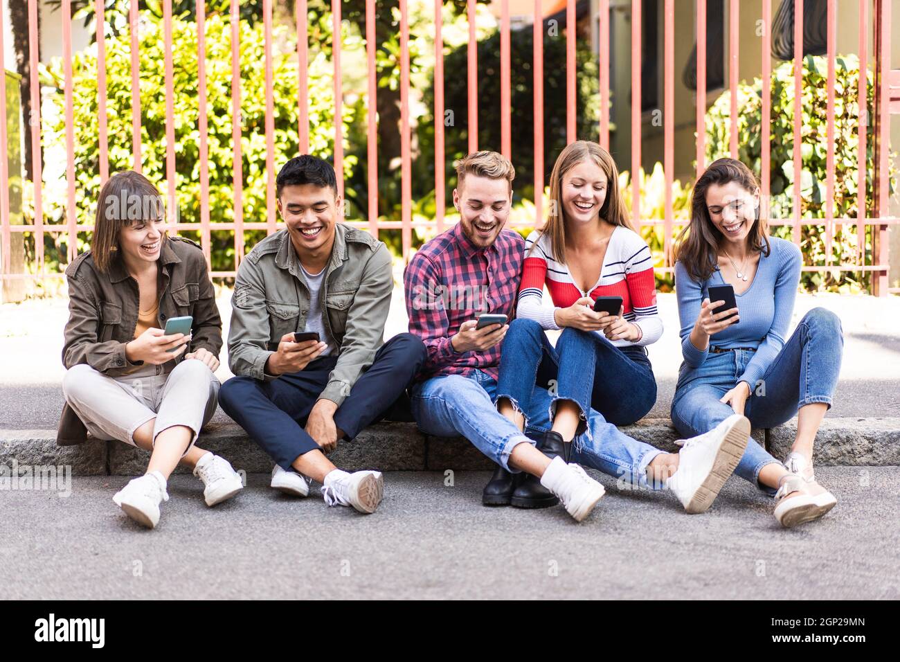 Des amis multiculturels riant en utilisant un smartphone assis sur le trottoir dans la ville - les jeunes toxicomanes par les téléphones mobiles intelligents - la technologie concep Banque D'Images