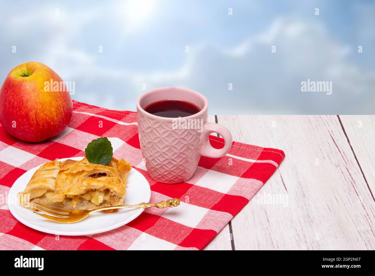 Rouleau de pommes servi. Une tranche de strudel aux pommes fraîches maison avec une tasse de thé et une pomme fraîche sur un tissu de pique-nique rouge sur fond clair. Conce Banque D'Images