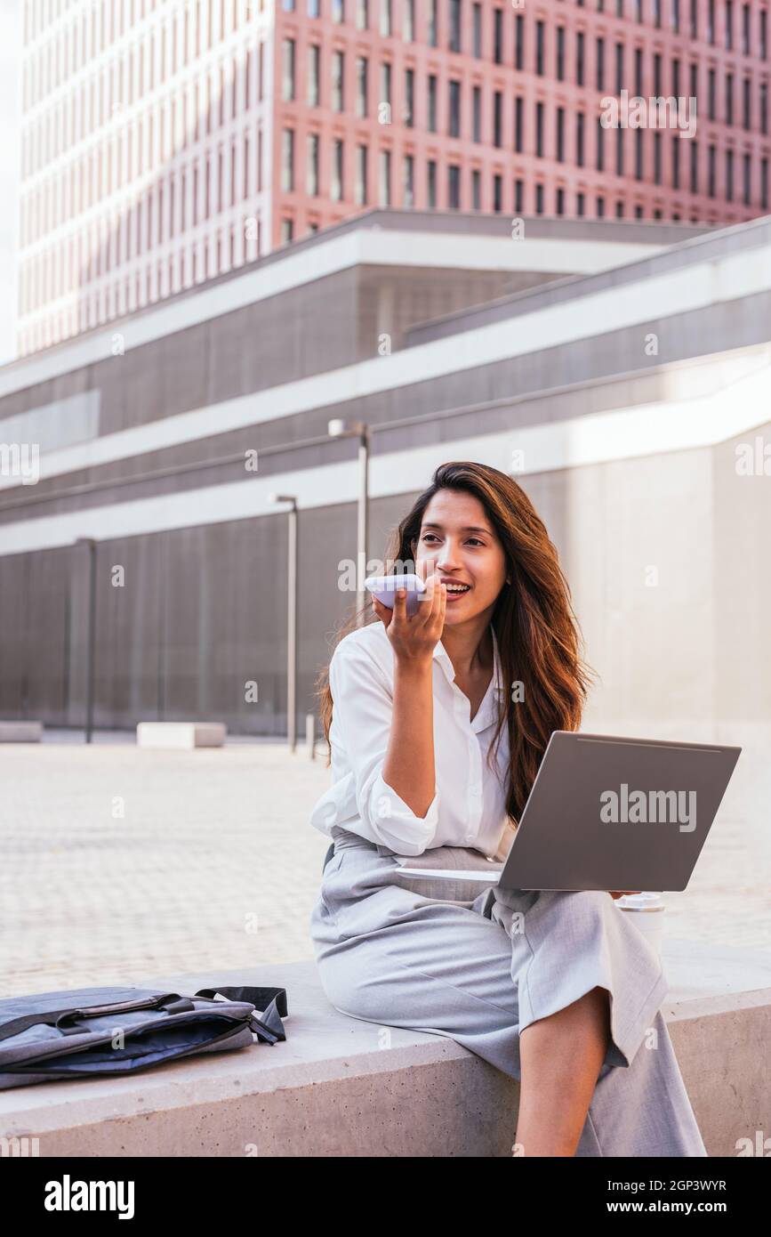 portrait vertical d'une jeune femme indienne élégante. Elle est assise près de l'immeuble de bureaux et laisse un message vocal avec le téléphone Banque D'Images