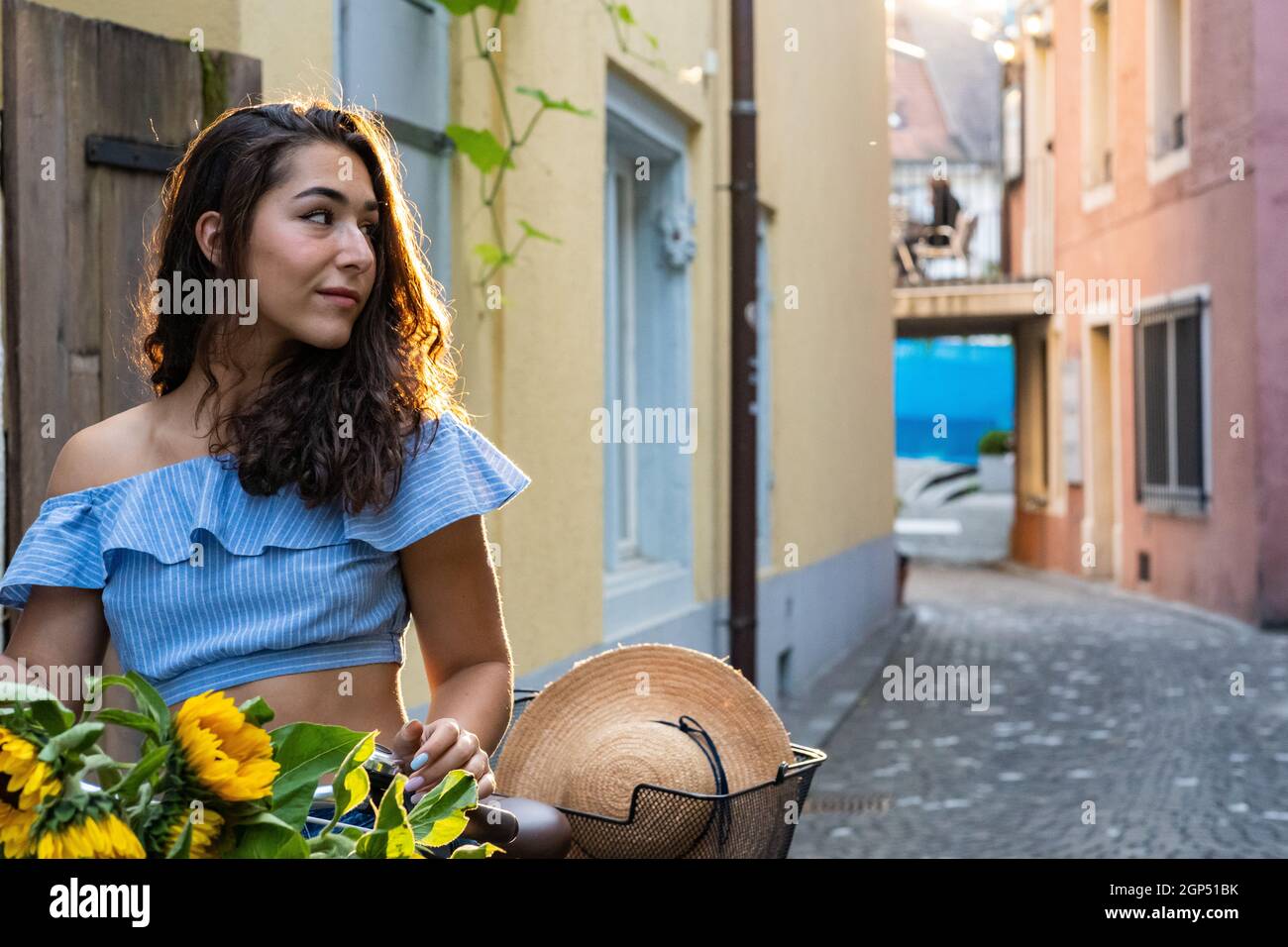 Belle jeune femme marchant dans la vieille ville tout en poussant son vélo turquoise avec des tournesols dans un panier. Sunbeam en arrière-plan. Banque D'Images