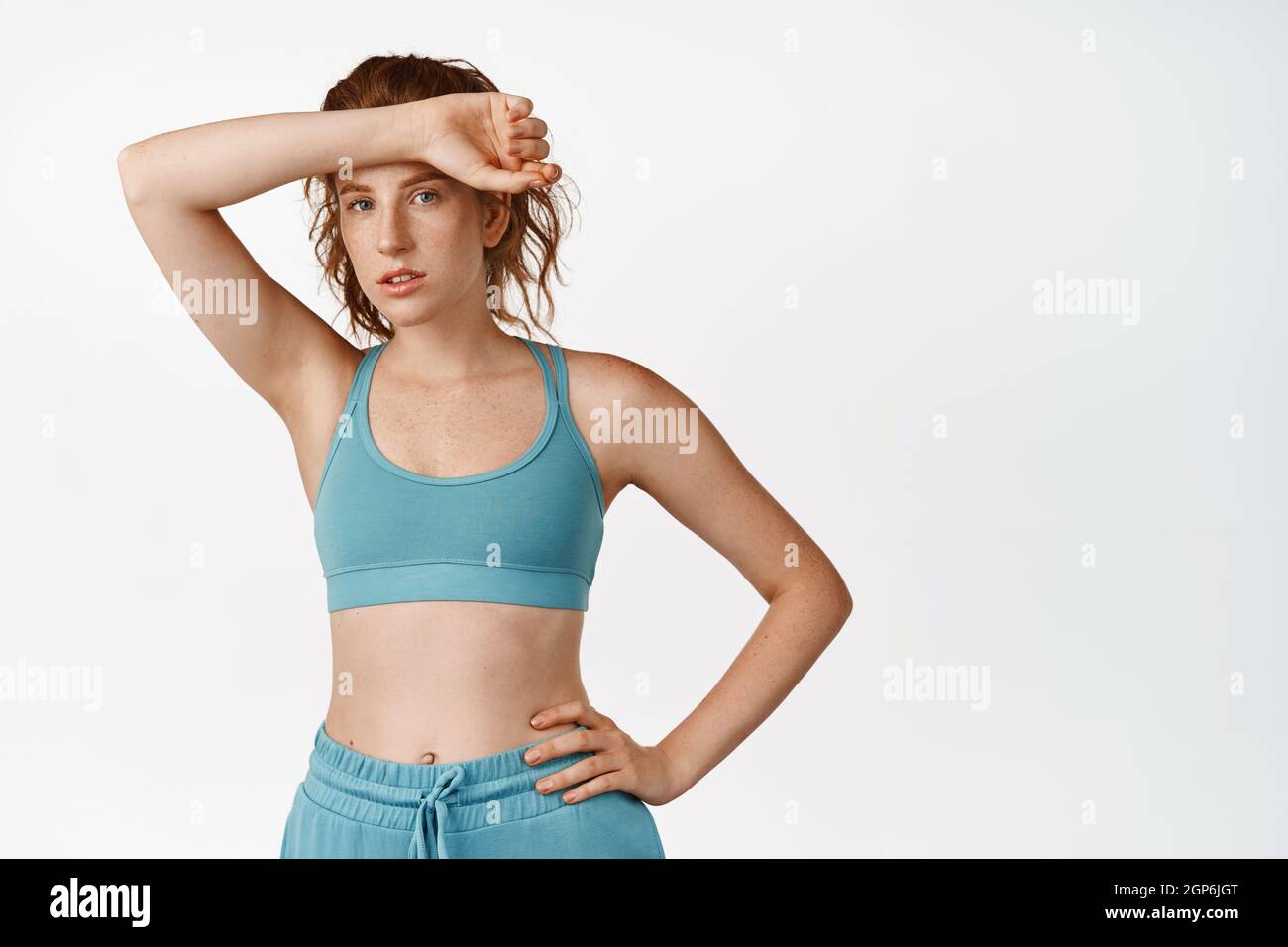 Jeune fille sportive dans des vêtements de fitness, essuyant la transpiration sur le front après séance d'entraînement, faisant du sport dans la salle de gym, debout sur fond blanc Banque D'Images