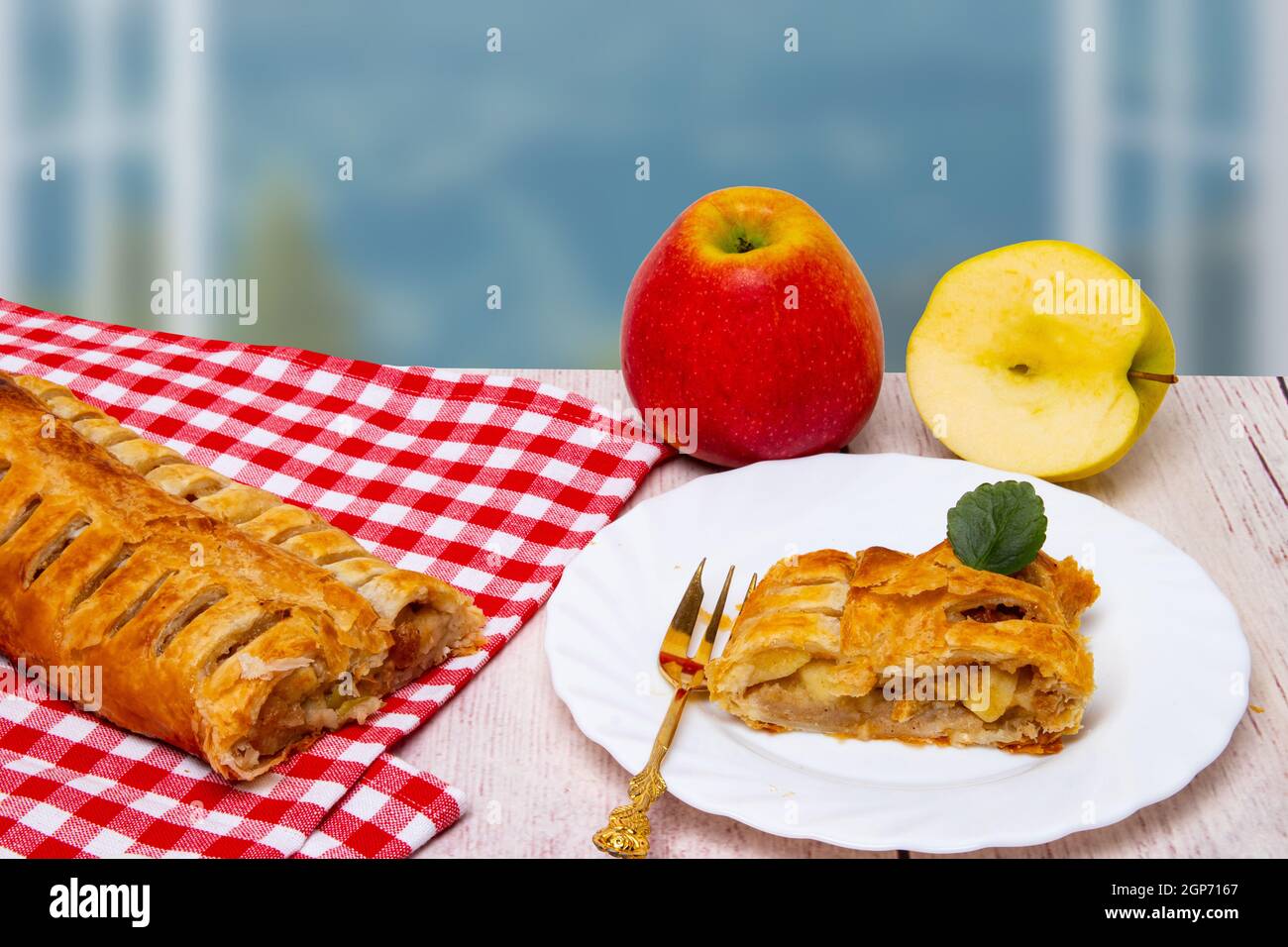 Un rouleau de strudel aux pommes fraîches maison avec une tranche appétissante de tarte aux pommes sur l'assiette et des pommes fraîches sur un fond de fenêtres ouvertes. Russe Banque D'Images