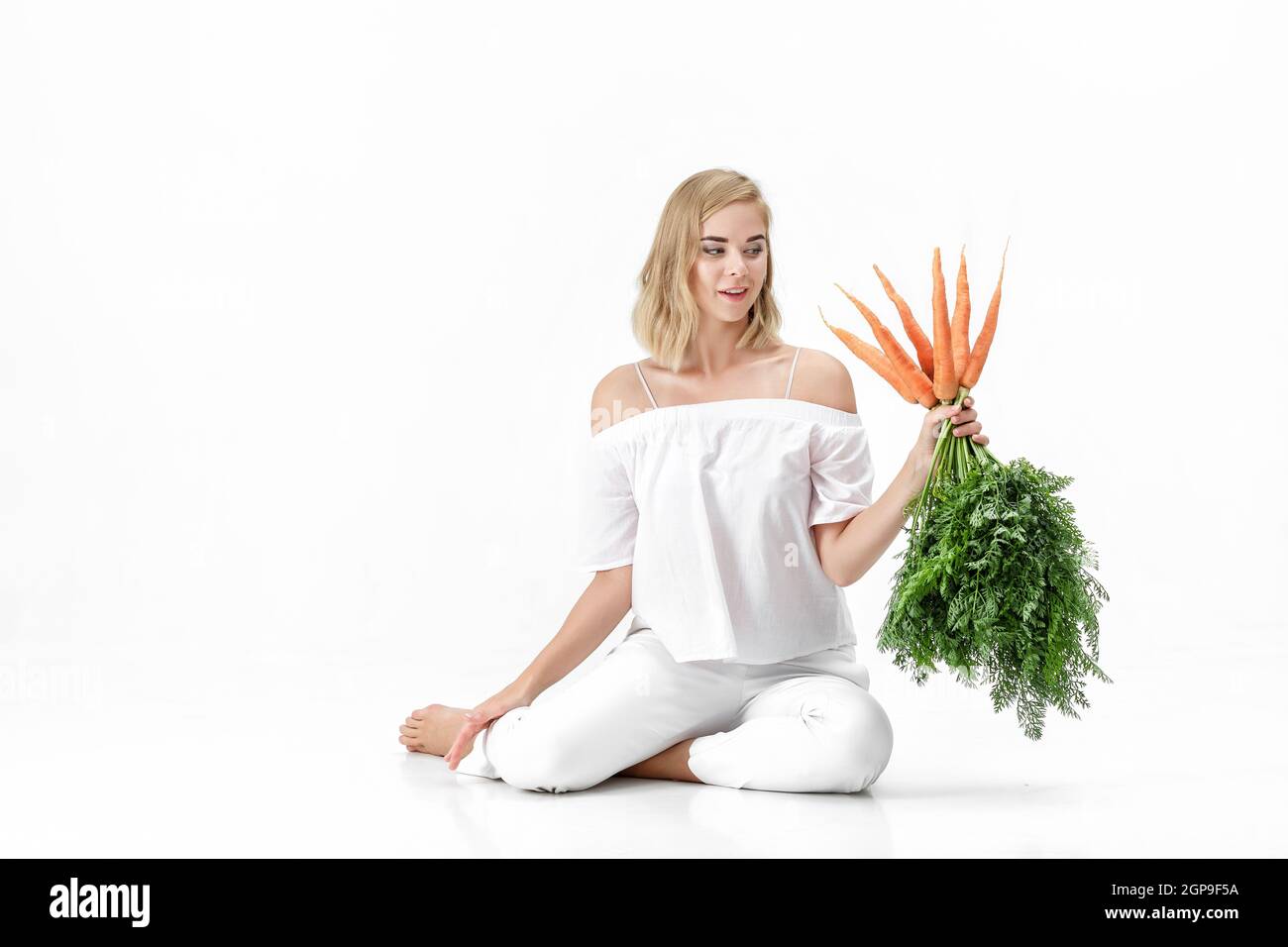 Belle femme blonde dans un chemisier blanc tenant une carotte fraîche avec des feuilles vertes sur un fond blanc. Santé et alimentation Banque D'Images