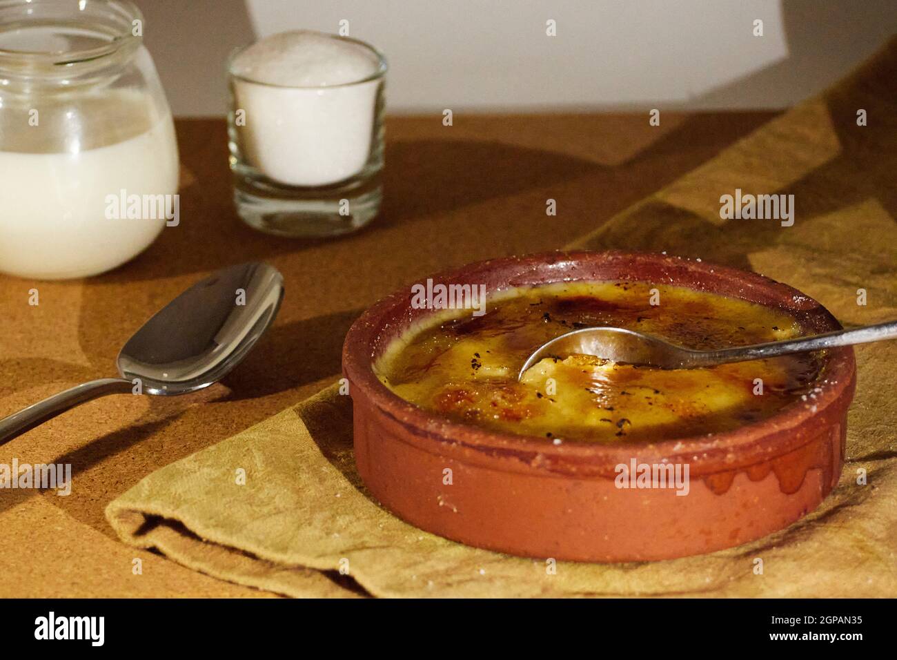 Cuillère plongée dans la crème catalane sur une serviette décorée d'une tasse de sucre et d'une tasse de lait. Sur la table en bois Banque D'Images