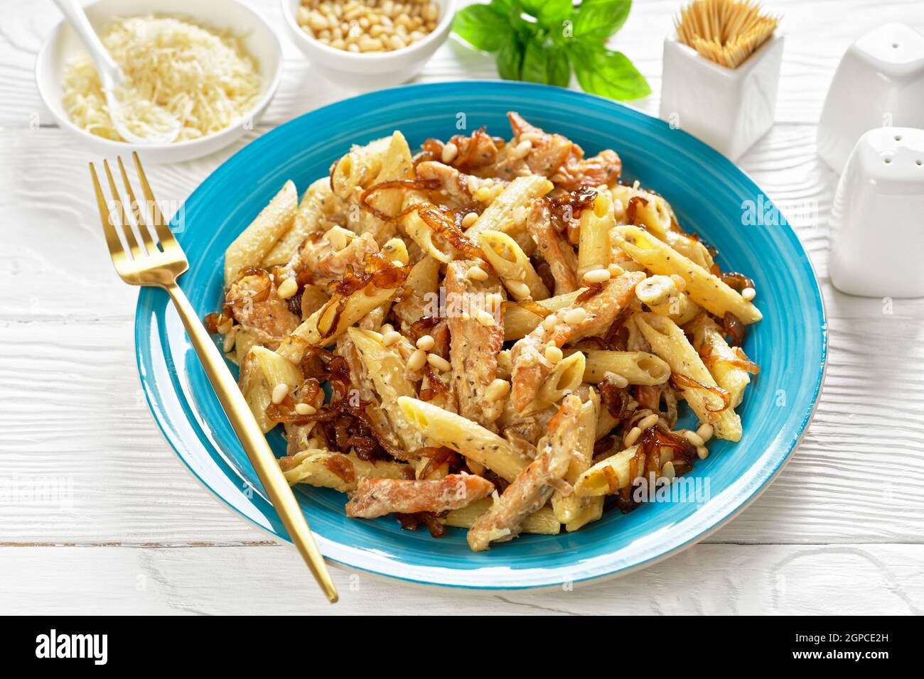 penne de pâtes alfredo de poulet à l'oignon caramélisé arrosé de pignons de pin et de parmesan râpé sur une assiette bleue sur une table en bois blanc Banque D'Images
