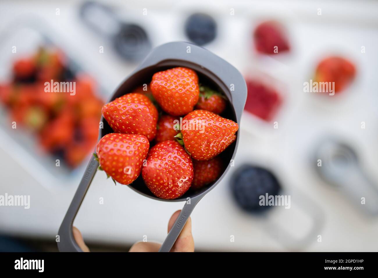 Petite tasse graduée pleine de fraises juteuses au-dessus de fruits plus flous. Baies fraîches et floues dans des récipients en plastique. Préparation de desserts fruités Banque D'Images