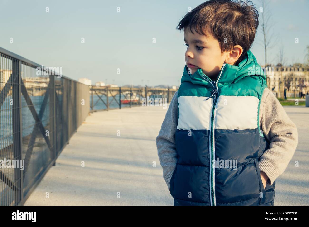 Portrait d'un petit garçon portant une veste sans manches, enfant regardant loin de l'appareil photo, se tient sur la promenande le long de la rivière. Banque D'Images