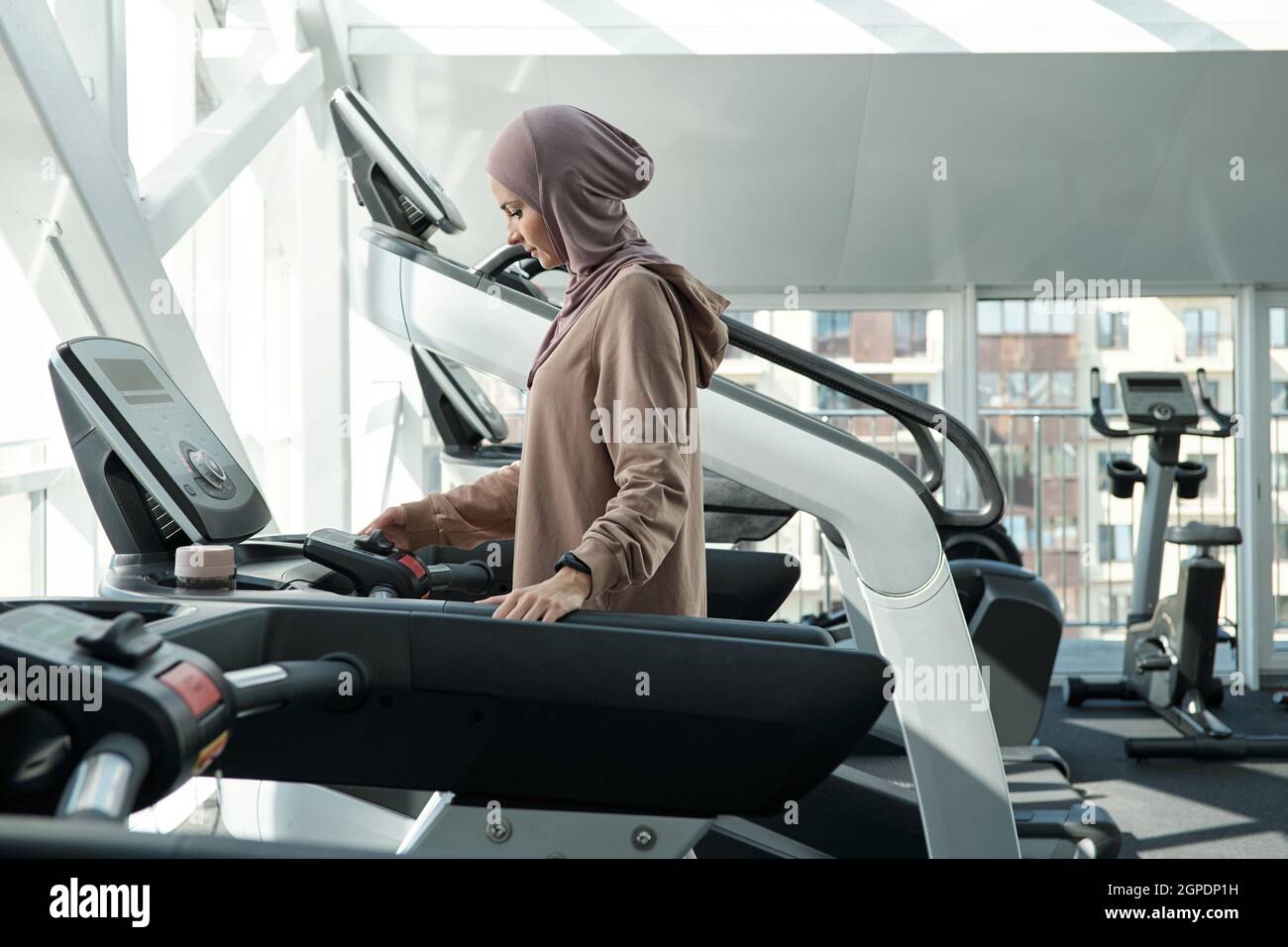 Vue latérale de la belle femme musulmane portant le hijab passant du temps dans la salle de gym debout sur le tapis roulant réglage des paramètres Banque D'Images