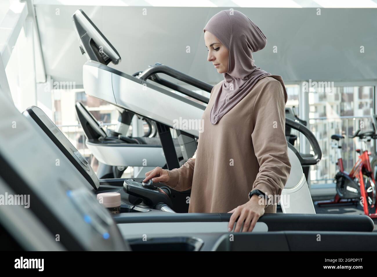 Vue latérale de la femme musulmane moderne portant le hijab en commençant le matin avec l'exercice dans la salle de gym debout sur le tapis roulant réglages Banque D'Images