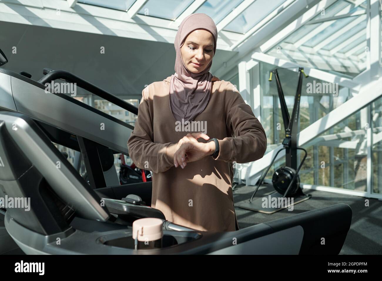 Portrait horizontal moyen d'une jeune femme musulmane adulte moderne portant le hijab debout sur un tapis roulant en salle de gym en vérifiant quelque chose sur son tracker d'activité Banque D'Images