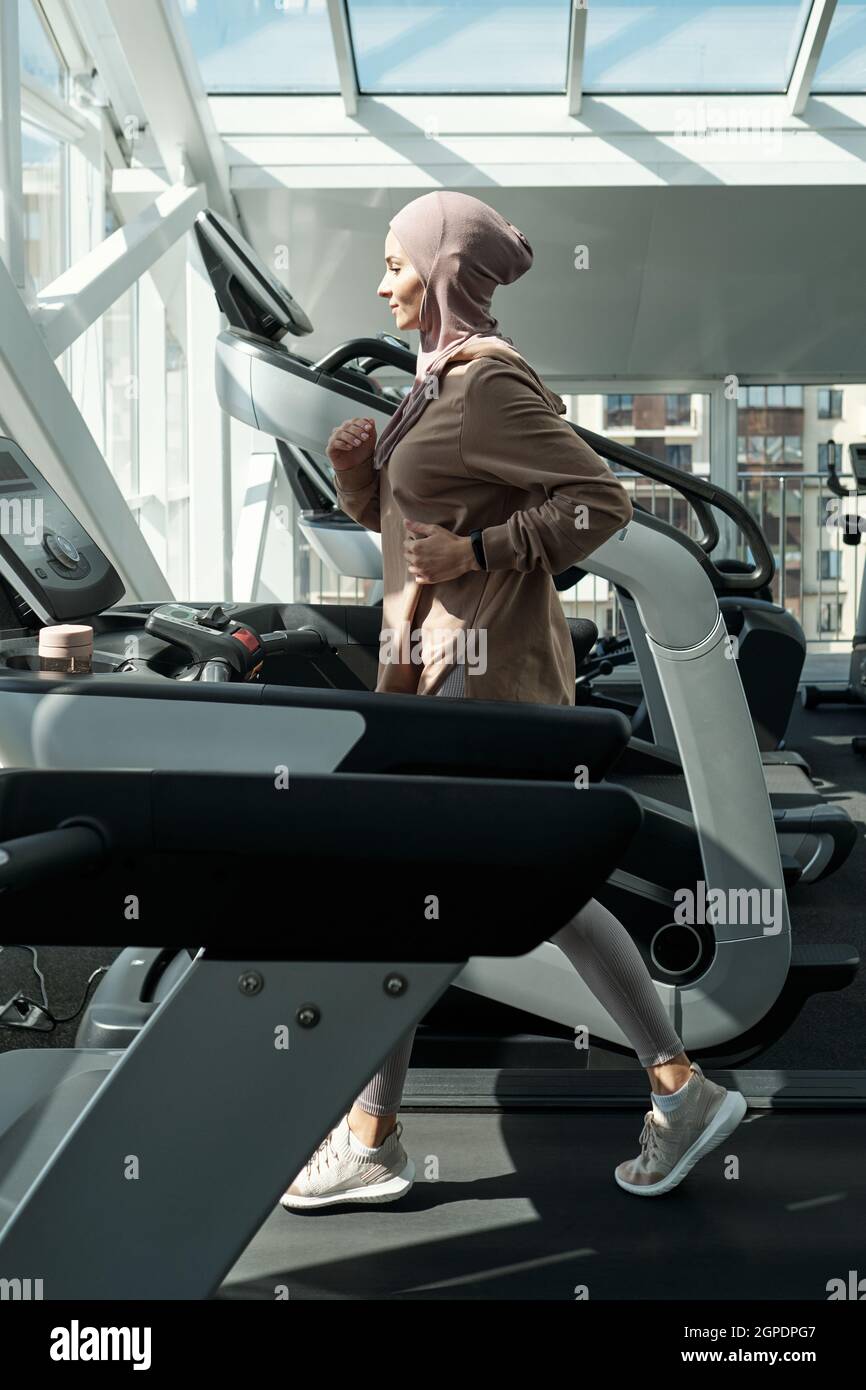 Vue latérale verticale image complète d'une femme musulmane sportive moderne portant le hijab en train de courir en salle de gym avec un tapis roulant Banque D'Images