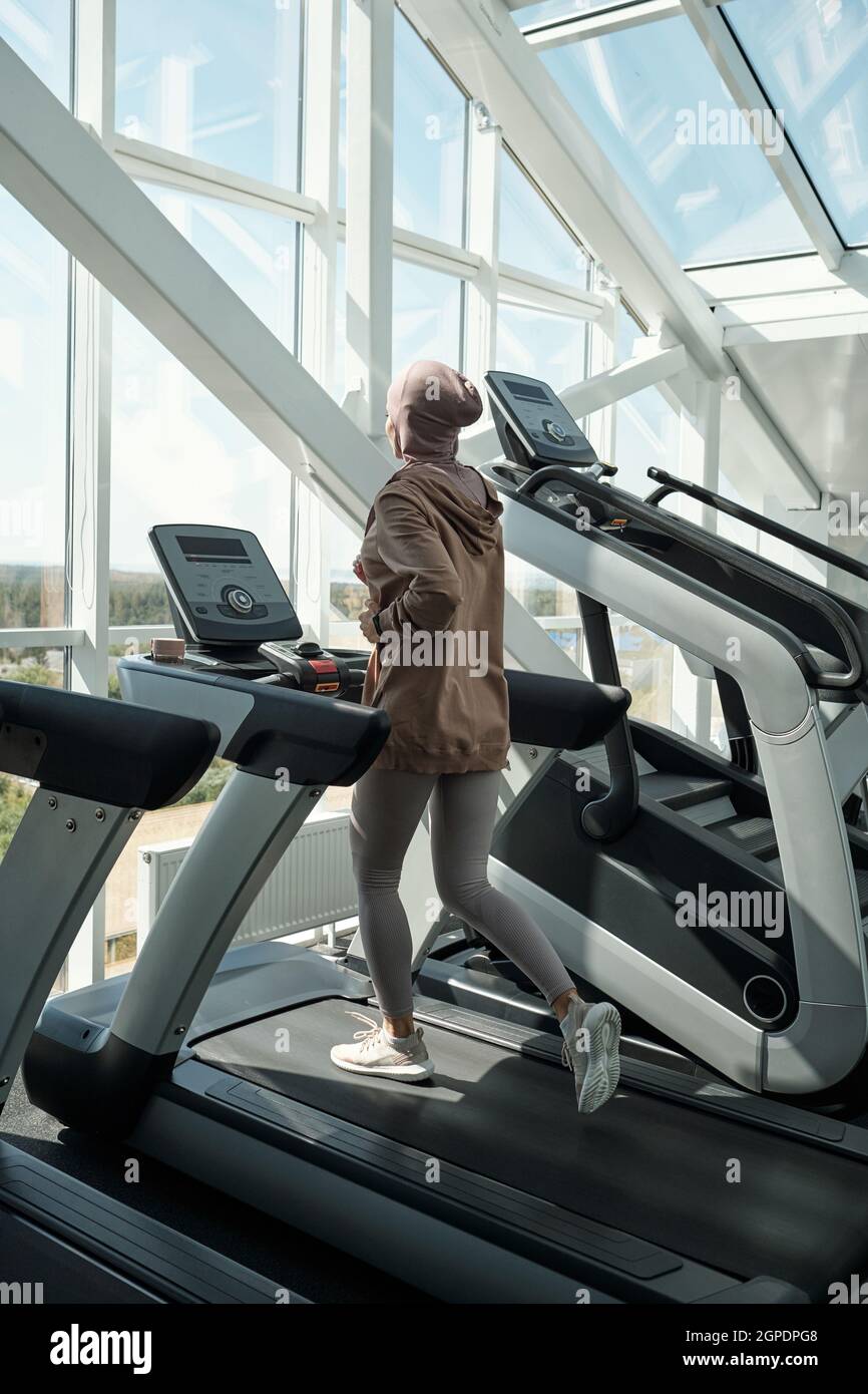 Plan vertical long de la jeune femme non reconnaissable portant hijab courir sur un tapis roulant machine d'exercice dans la salle de gym moderne Banque D'Images