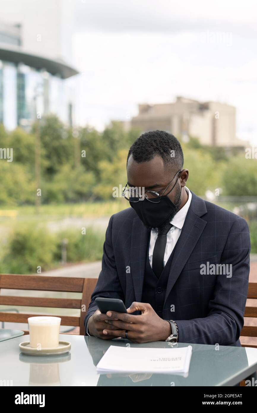 Jeune homme d'affaires africain en costume et masque noir textant dans un smartphone par table dans un café extérieur Banque D'Images