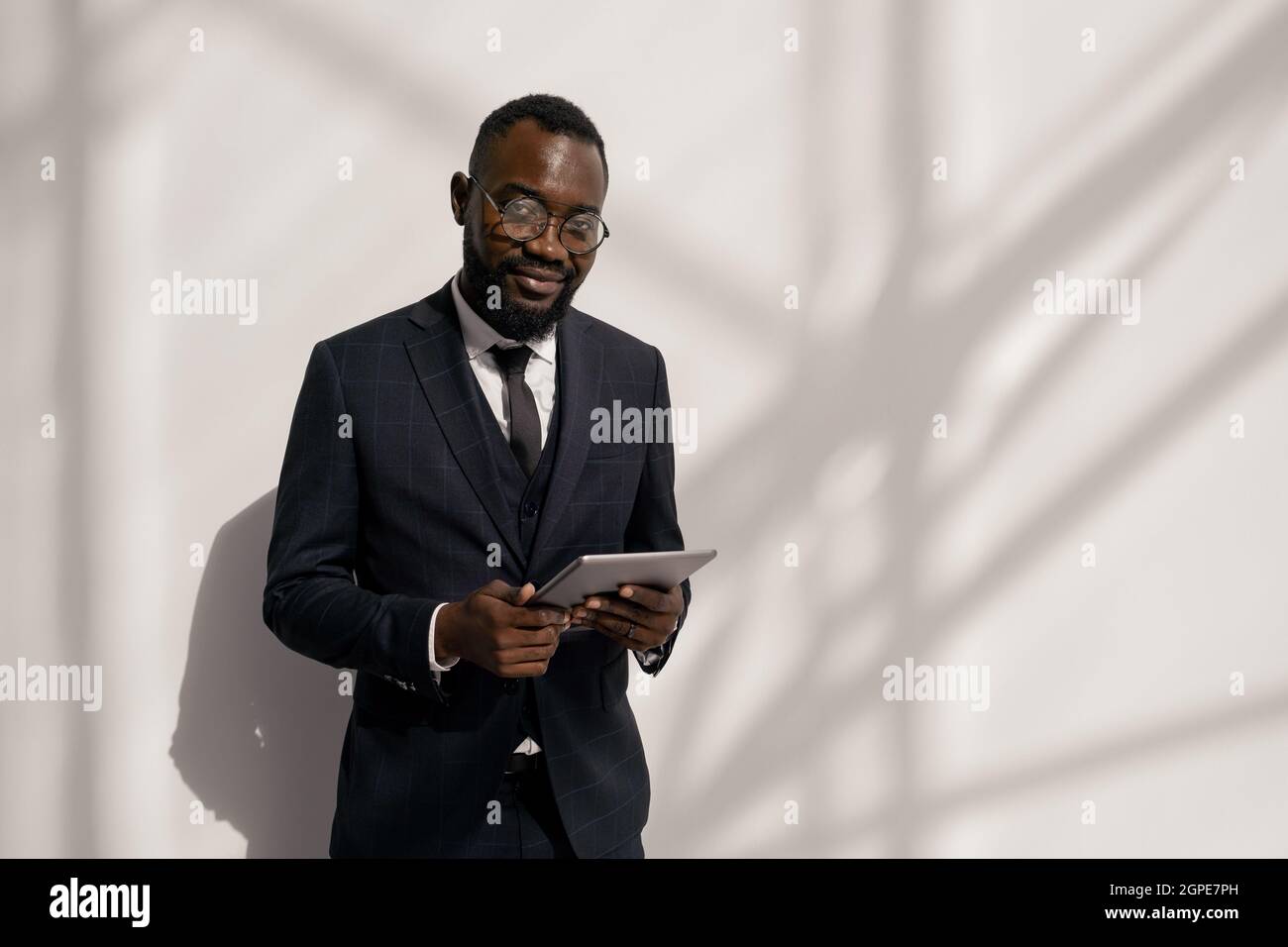 Joyeux jeune Manager africain en costume défilant dans la tablette contre le mur blanc Banque D'Images