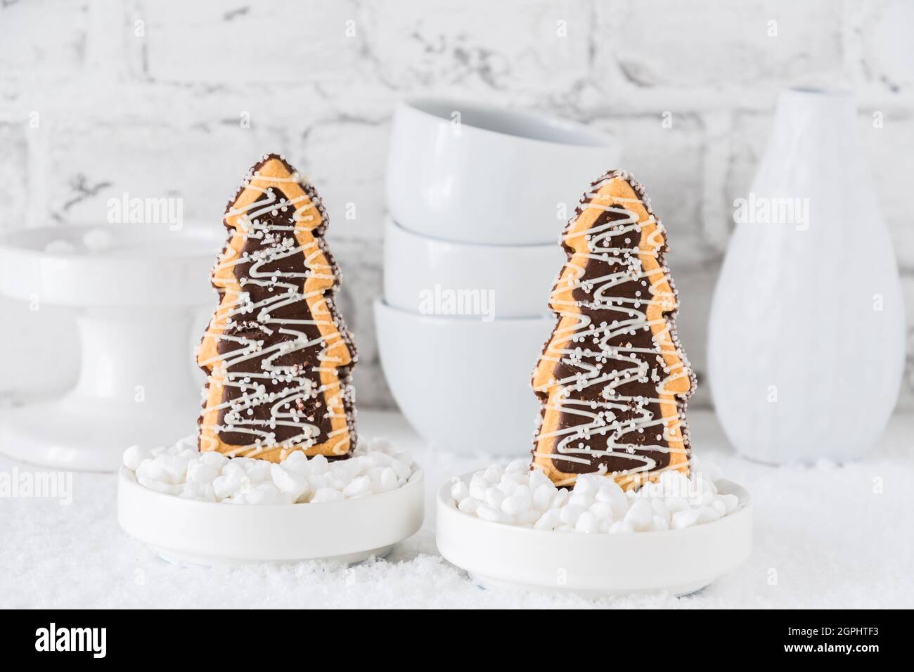 Deux biscuits en forme d'arbres de noël avec chocolat et sucre glacer dans des bols avec du sucre grossier sur fond blanc avec de la vaisselle blanche Banque D'Images