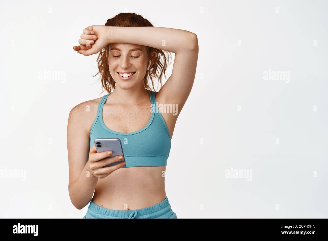 Une jeune fille en forme souriante se satisfait après l'entraînement, essuyant la transpiration du front et regardant l'application sport pour smartphone, se tenant sur un fond blanc Banque D'Images