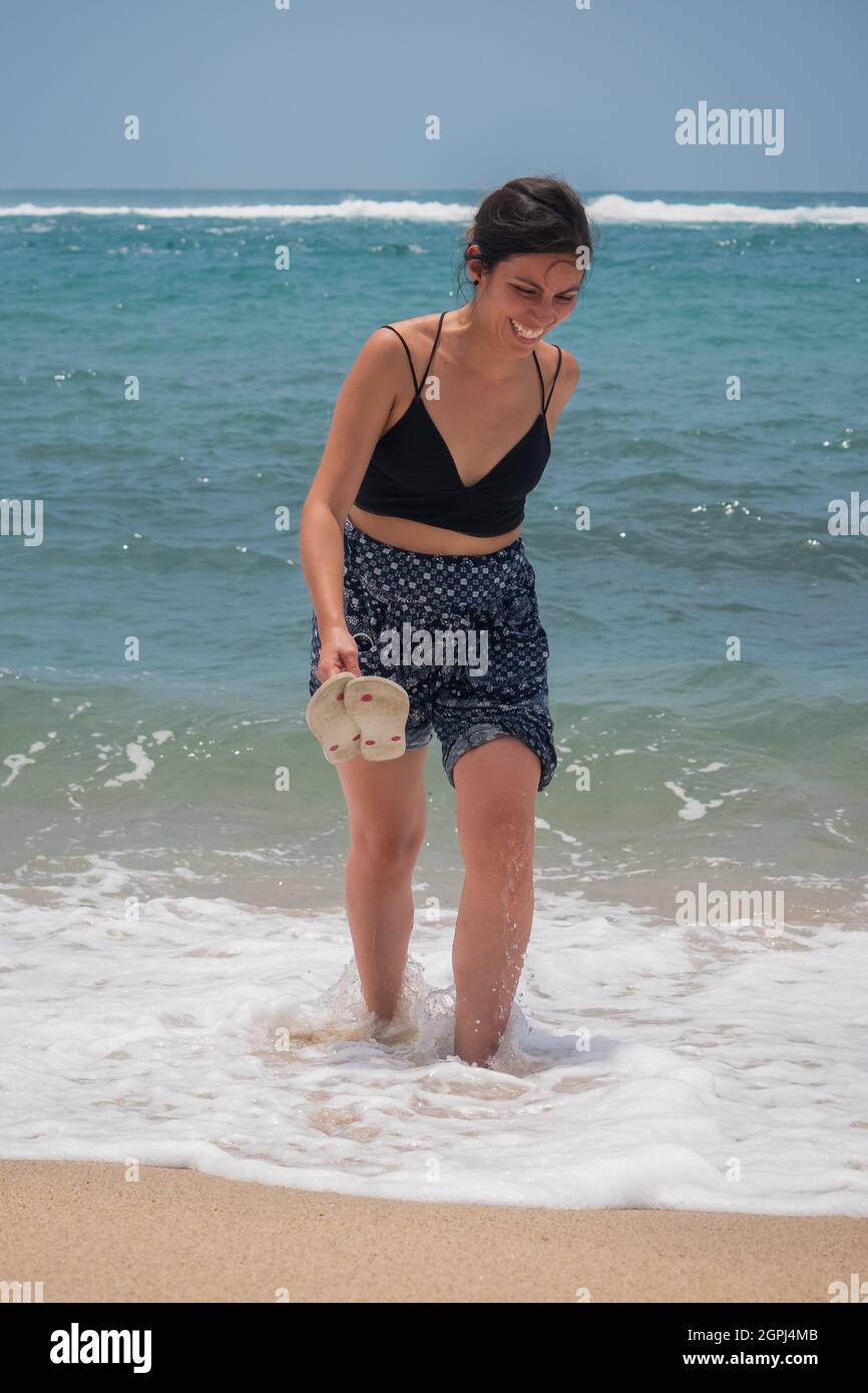 Jeune Latina tenant une paire de sandales et marchant le long de la mer riant beaucoup Banque D'Images
