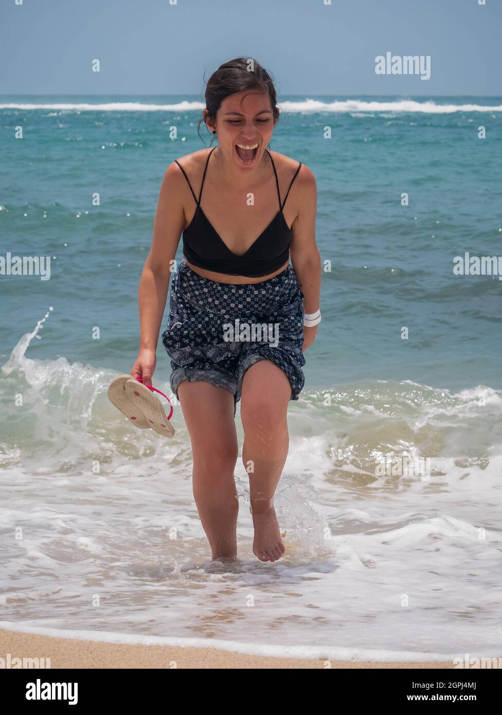 Jeune Latina tenant une paire de sandales et marchant le long de la mer riant beaucoup Banque D'Images