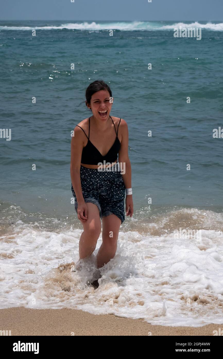 Jeune Latina tenant une paire de sandales et marchant le long de la mer riant beaucoup Banque D'Images