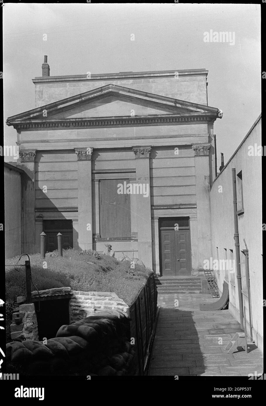 Chapelle à Hull Trinity House et statue à l'extérieur du front est, Princes Dock Street, Kingston upon Hull, 1941. Vue extérieure de l'élévation est de la chapelle de la Maison de la Trinité, avec vue partielle sur une sculpture d'Oceanus et un abri à la bombe au premier plan. La chapelle date de 1842 et fut construite par H. F. Lockwood. L'élévation est est rustiquée et a quatre pilastres corinthiens géants soutenant une frise et un pediment, avec la fenêtre est au centre. La fenêtre est entourée de deux portes à huit panneaux avec pilastres en pierre et lampes rectangulaires. En face de la fenêtre est se trouve a s. Banque D'Images