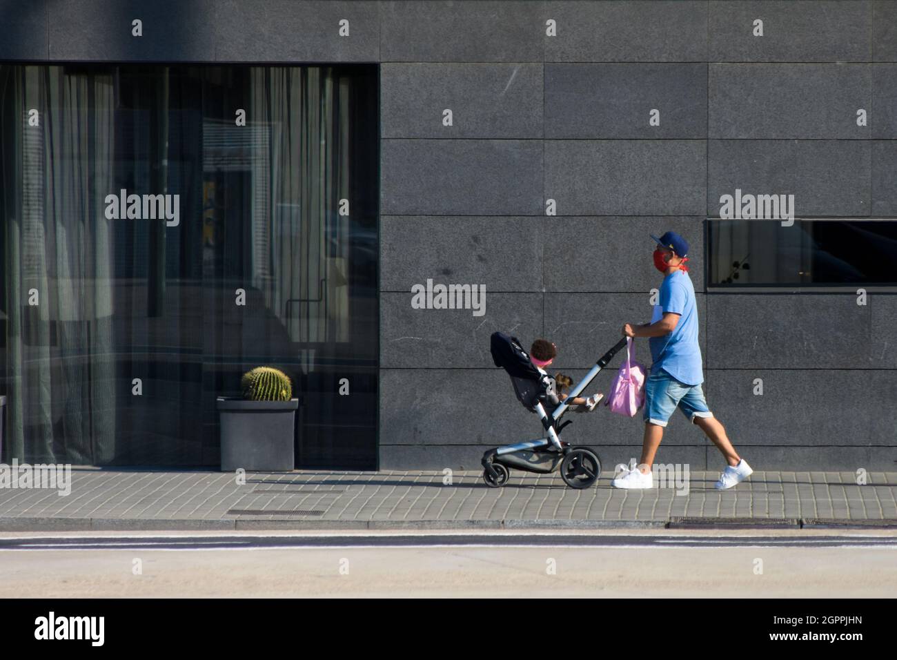 Père avec masque poussant son bébé dans une poussette dans une rue près d'un mur. Banque D'Images