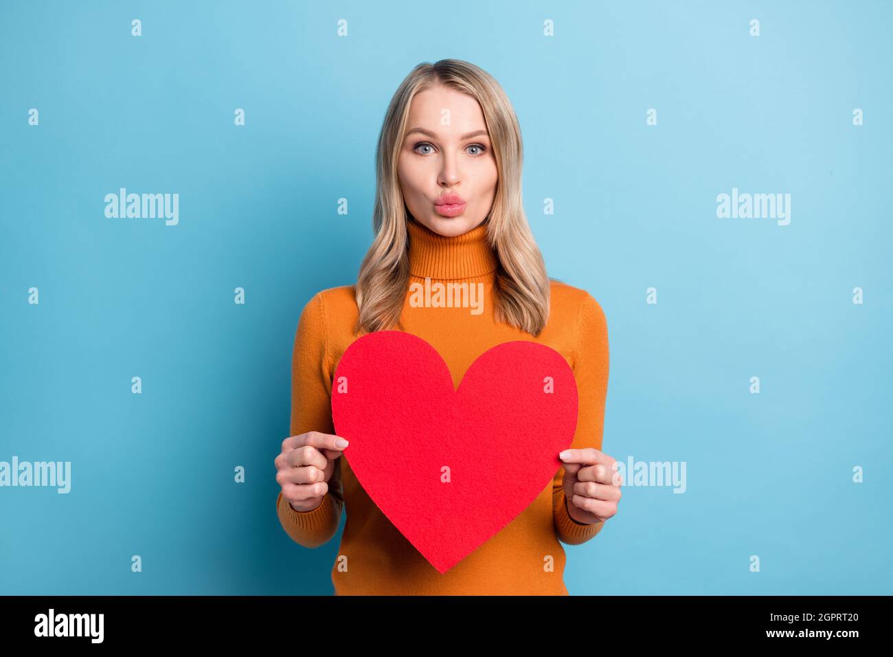 Photo de brillant flirty jeune femme porter un chandail orange tenant le grand coeur vous envoyant baiser isolé couleur bleu arrière-plan Banque D'Images