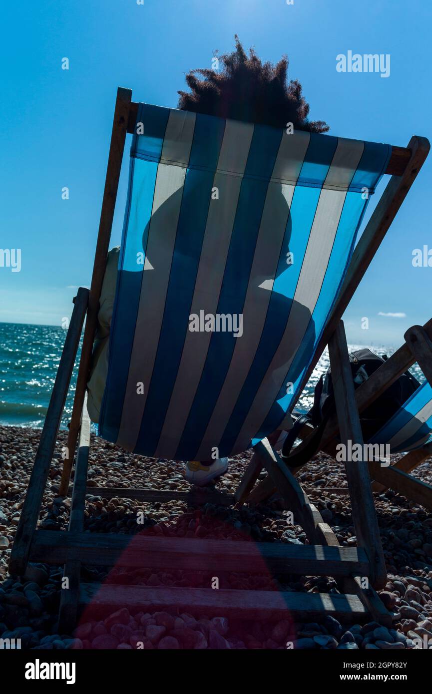 jeune femme se relaxant sur la plage dans un transat sous le soleil d'été.Sa coiffure afro est visible au-dessus de l'arrière du transat rayé Banque D'Images