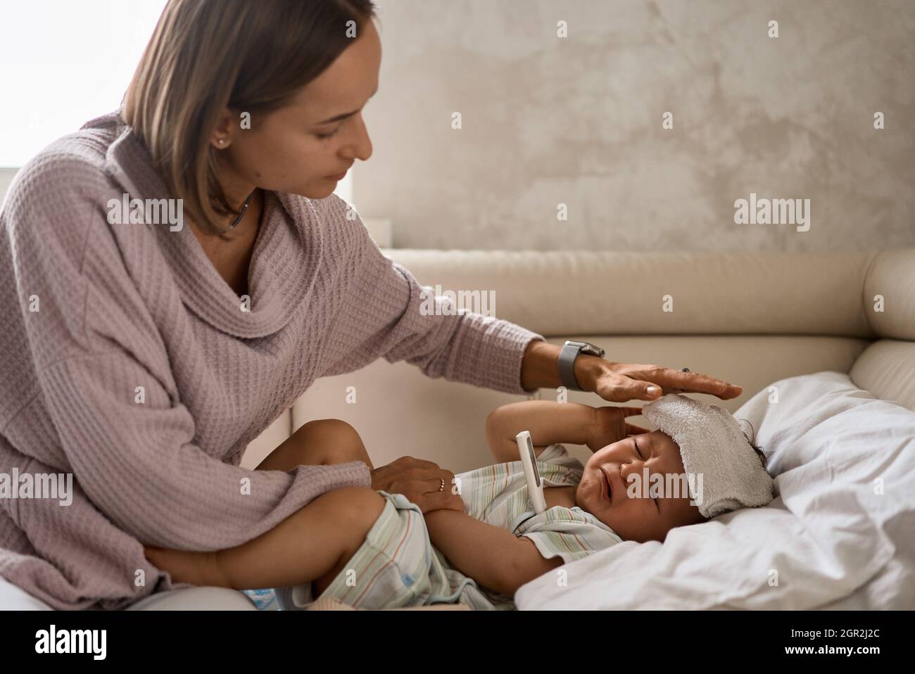 Jeune petit garçon sur le canapé avec gel froid sur sa tête et mesure de la température par thermomètre. Et prendre soin de sa mère Banque D'Images