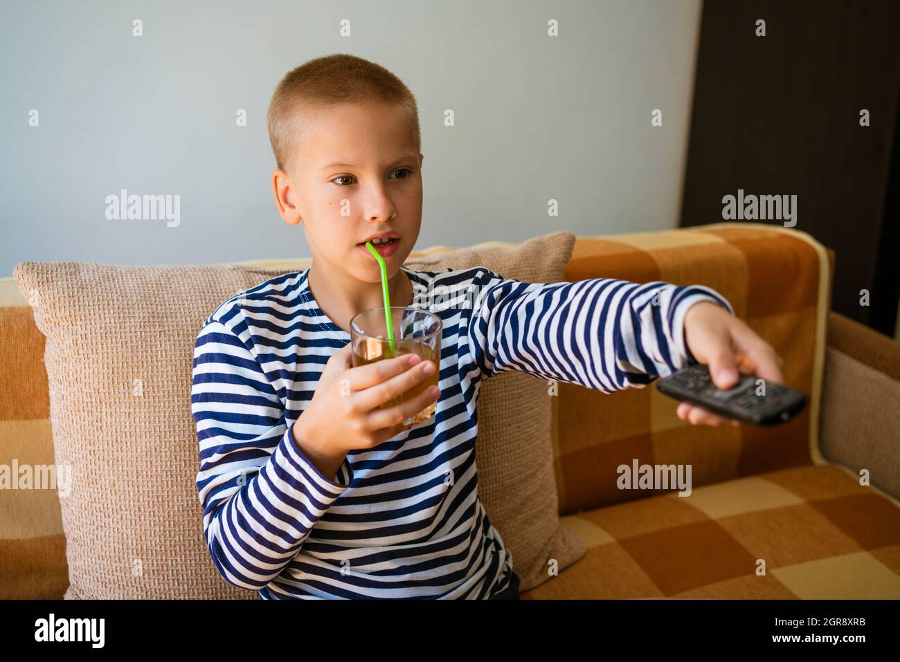 Un garçon d'âge scolaire caucasien est assis sur un canapé dans des vêtements décontractés avec une télécommande dans sa main et change de chaîne sur la télévision. Et boire du jus dans un verre à travers une paille. Concept de loisirs maison de vacances Banque D'Images