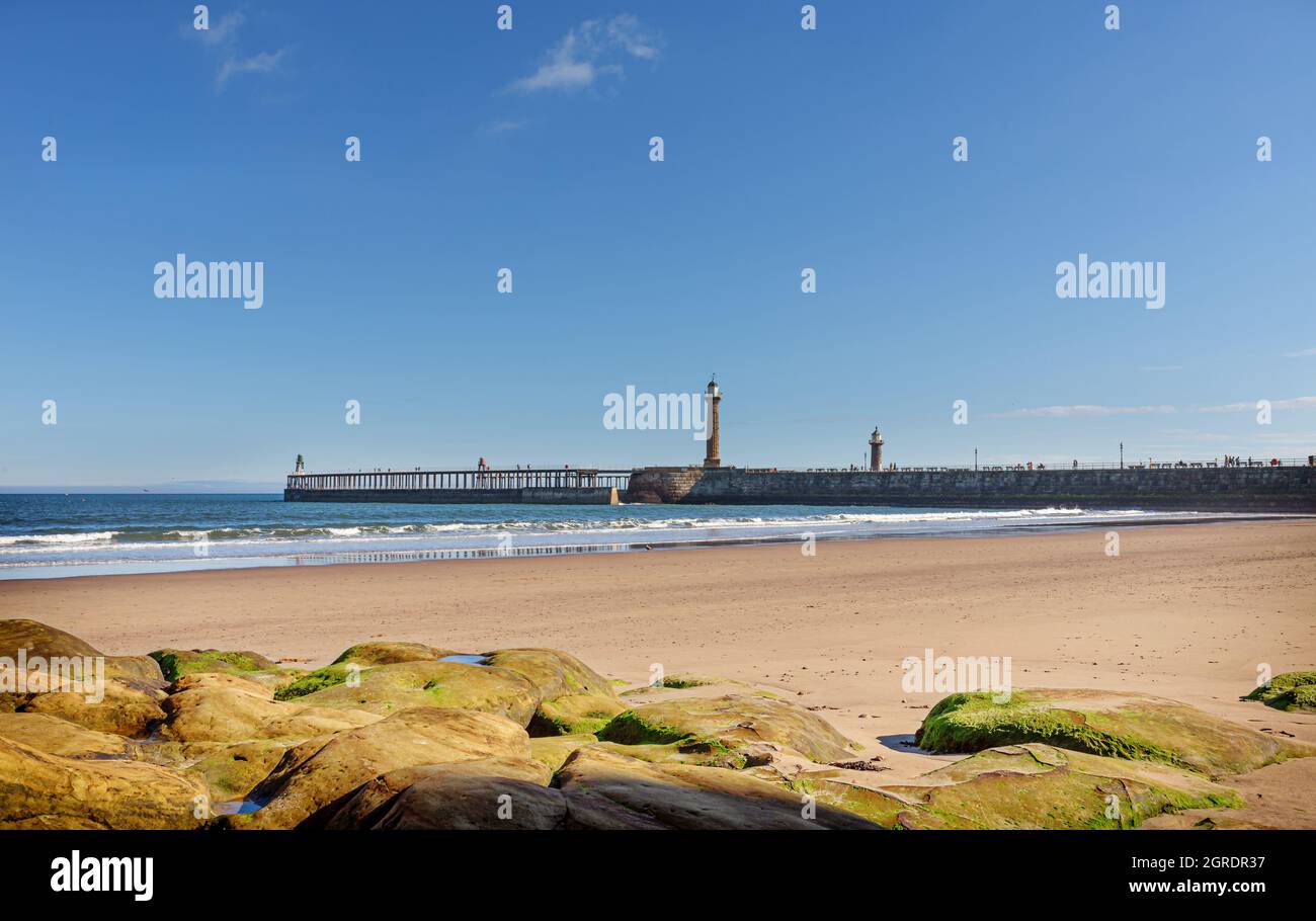 Une plage à marée basse avec des rochers au premier plan. Un long quai avec un phare s'étend jusqu'à la mer et un ciel avec un nuage léger est au-dessus. Banque D'Images