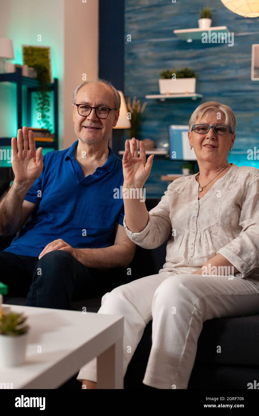 POV de couple à la retraite en agitant la main sur la vidéoconférence dans le salon. Personnes âgées regardant la caméra en étant assis sur un canapé et en utilisant la communication à distance en ligne. Vieux mari et femme Banque D'Images