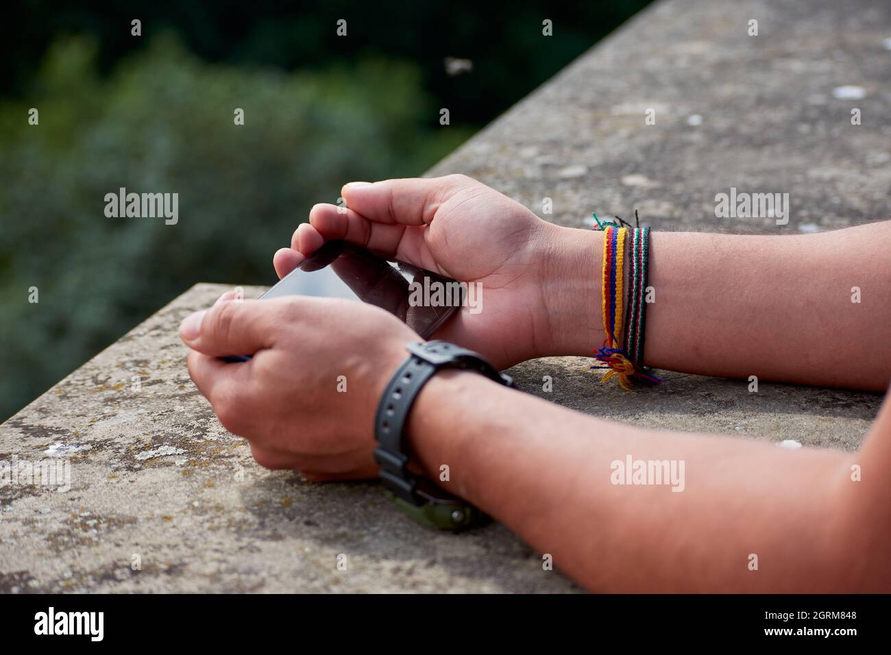Les mains des hommes portant une montre et des bracelets tenant un smartphone appuyé sur une structure en pierre Banque D'Images
