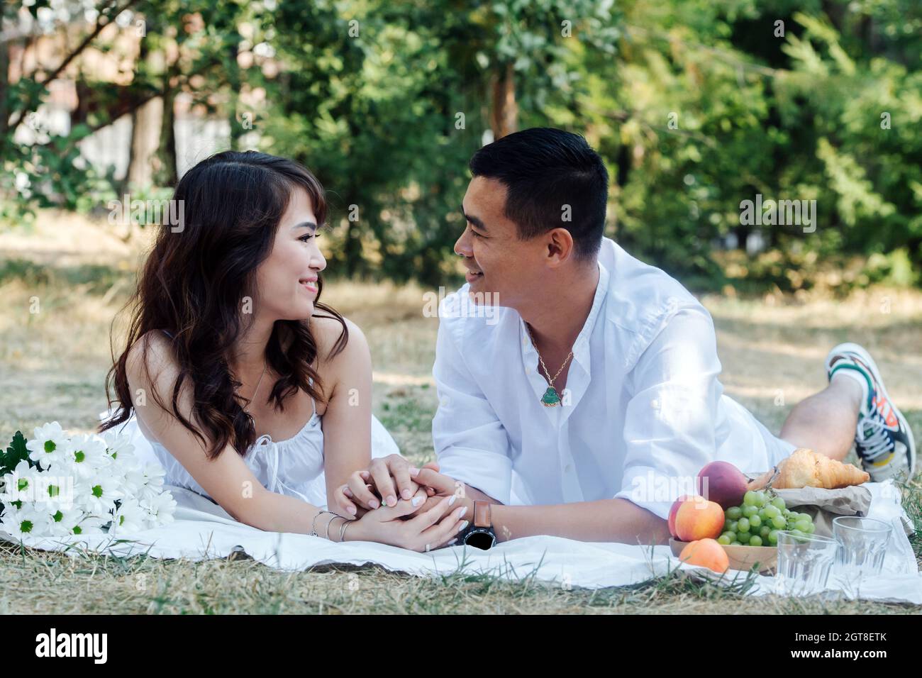 Couple asiatique aimant se happant pique-nique dans un parc, allongé sur leurs estomacs, regardant l'un l'autre. Homme et femme se reposant dans des vêtements blancs à l'ombre d'un arbre. Banque D'Images