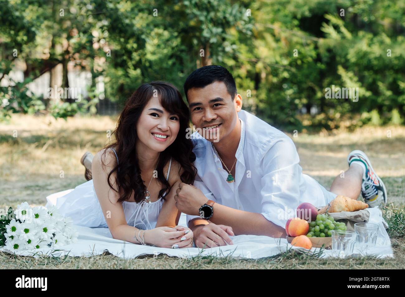 Un couple asiatique souriant se fait un pique-nique dans un parc, allongé sur leurs estomacs. Homme et femme en vêtements blancs dans une ombre d'arbre. Banque D'Images