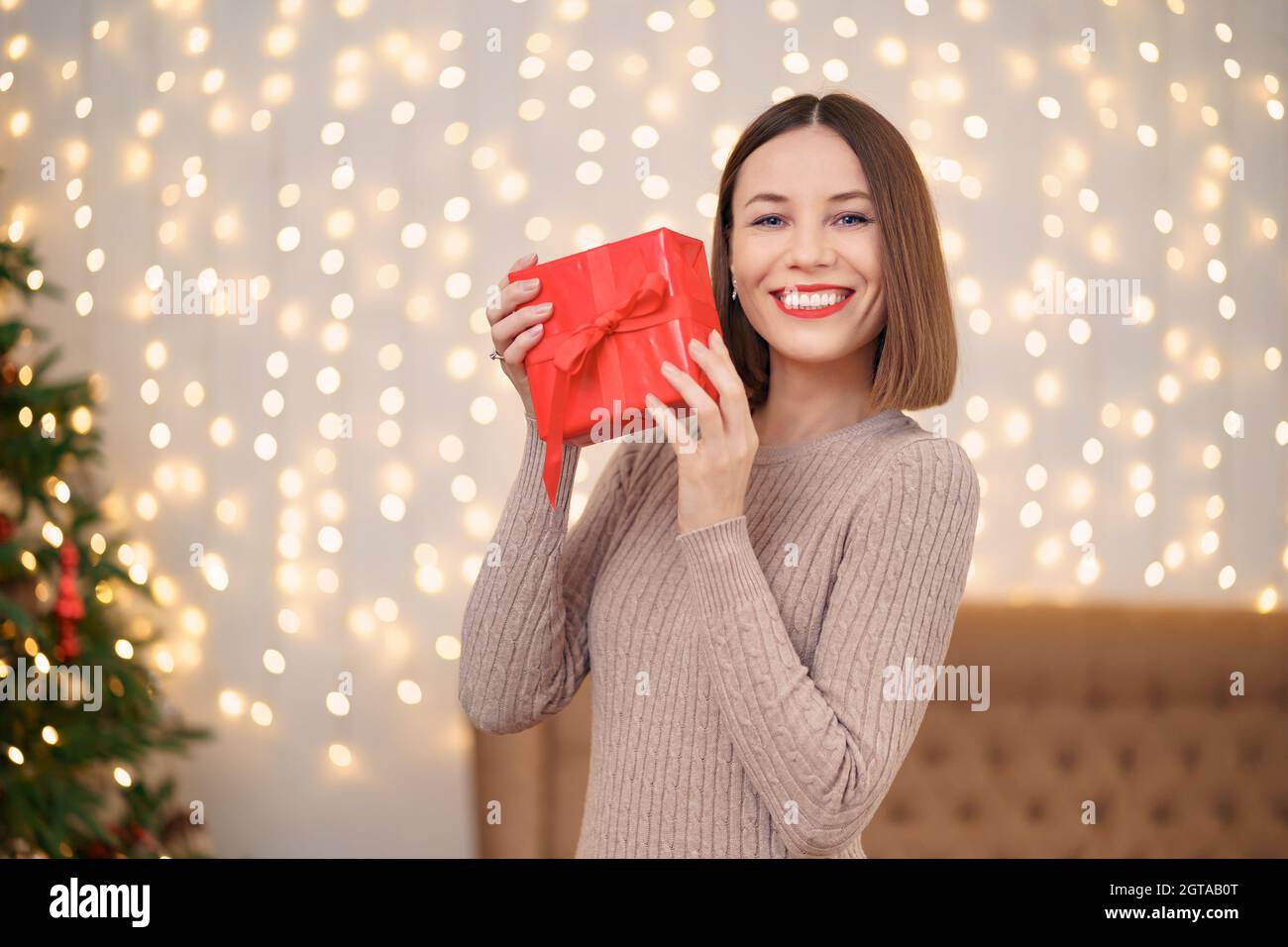 Portrait de jeune femme heureuse lèvres rouges regardant la boîte cadeau emballée.Gros plan la femme satisfaite a reçu la boîte actuelle.Arrière-plan des lumières de Noël festives. Banque D'Images