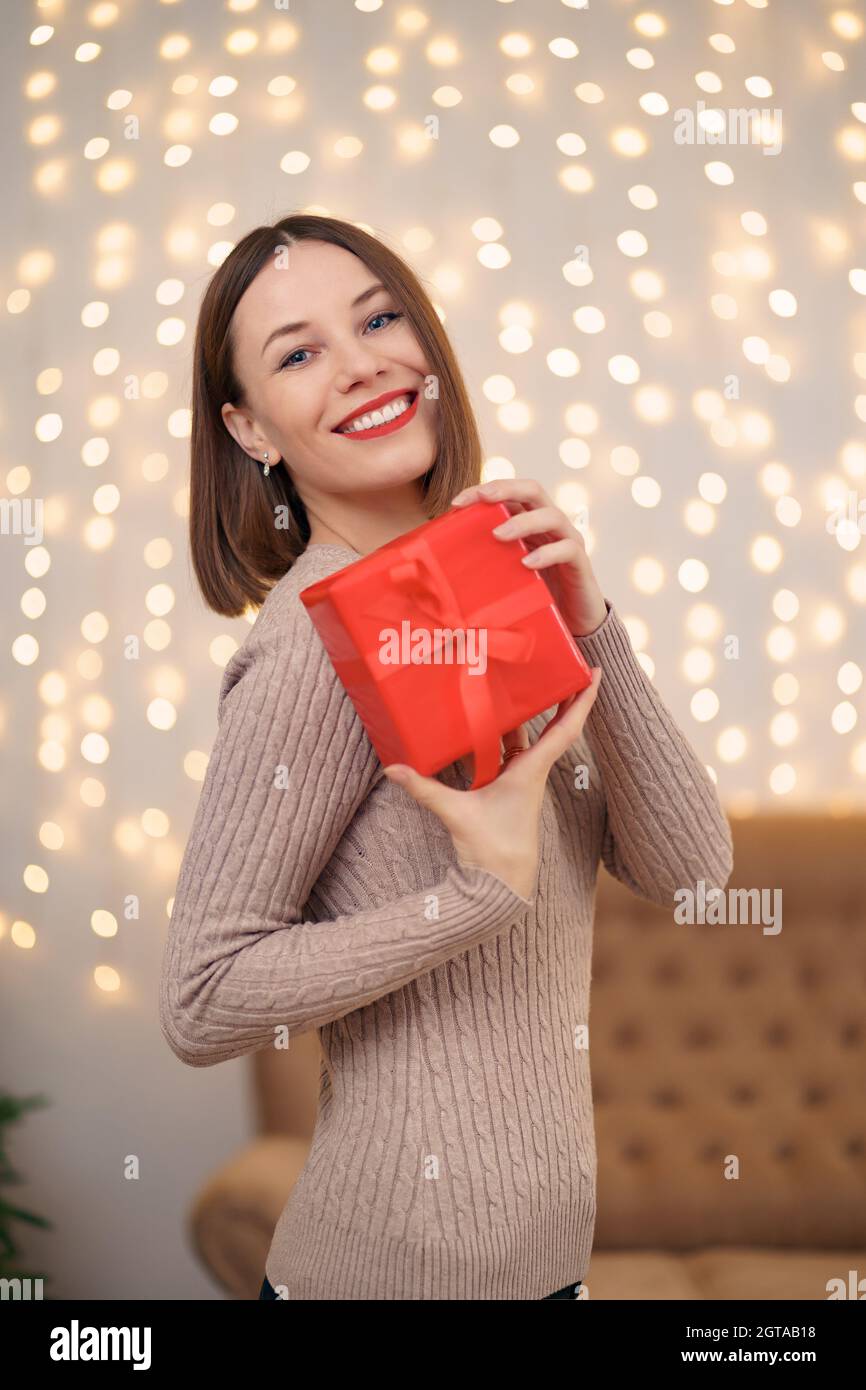 Portrait de jeune femme heureuse lèvres rouges posant avec une boîte cadeau emballée.Gros plan la femme satisfaite a reçu la boîte actuelle.Arrière-plan des lumières de Noël festives. Banque D'Images