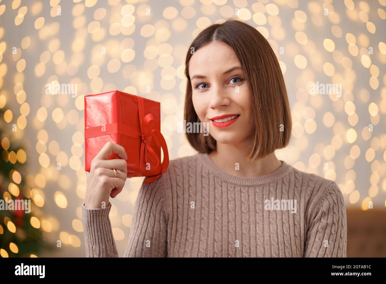 Portrait d'une jeune femme heureuse lèvres rouges tenant une boîte cadeau emballée. Gros plan la femme satisfaite a reçu la boîte actuelle. Arrière-plan des lumières de Noël festives. Banque D'Images