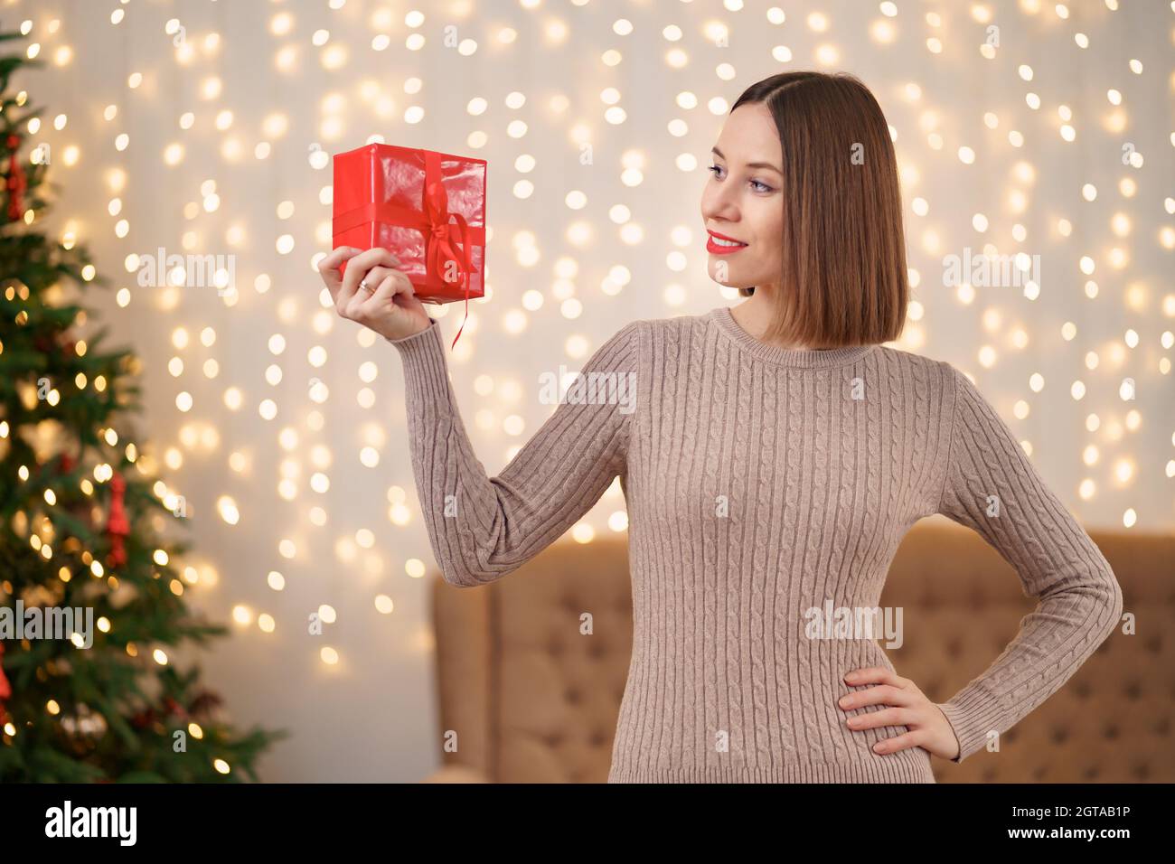 Portrait de jeune femme heureuse lèvres rouges regardant la boîte cadeau emballée.Gros plan la femme satisfaite a reçu la boîte actuelle.Arrière-plan des lumières de Noël festives. Banque D'Images