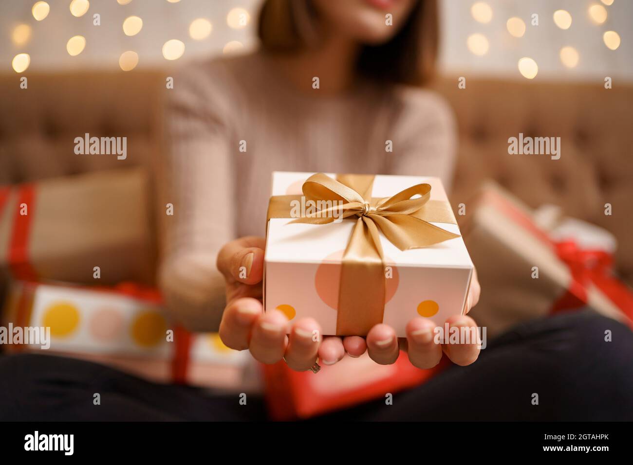 Une jeune femme heureuse tenant un cadeau de présentation entouré de cadeau des boîtes sont assises sur un canapé à dos de chameau avec des lumières en arrière-plan Banque D'Images
