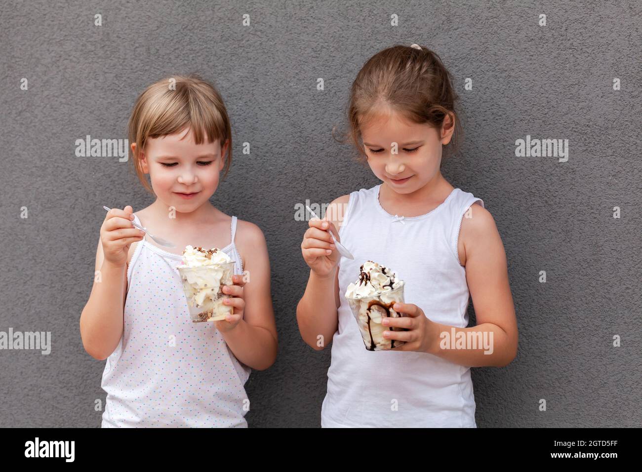 Deux jeunes filles gaies, sœurs, frères et sœurs, enfants mangeant de la crème glacée dans des tasses ensemble souriant, en utilisant des cuillères, portrait, gros plan. Enfants et doux Banque D'Images