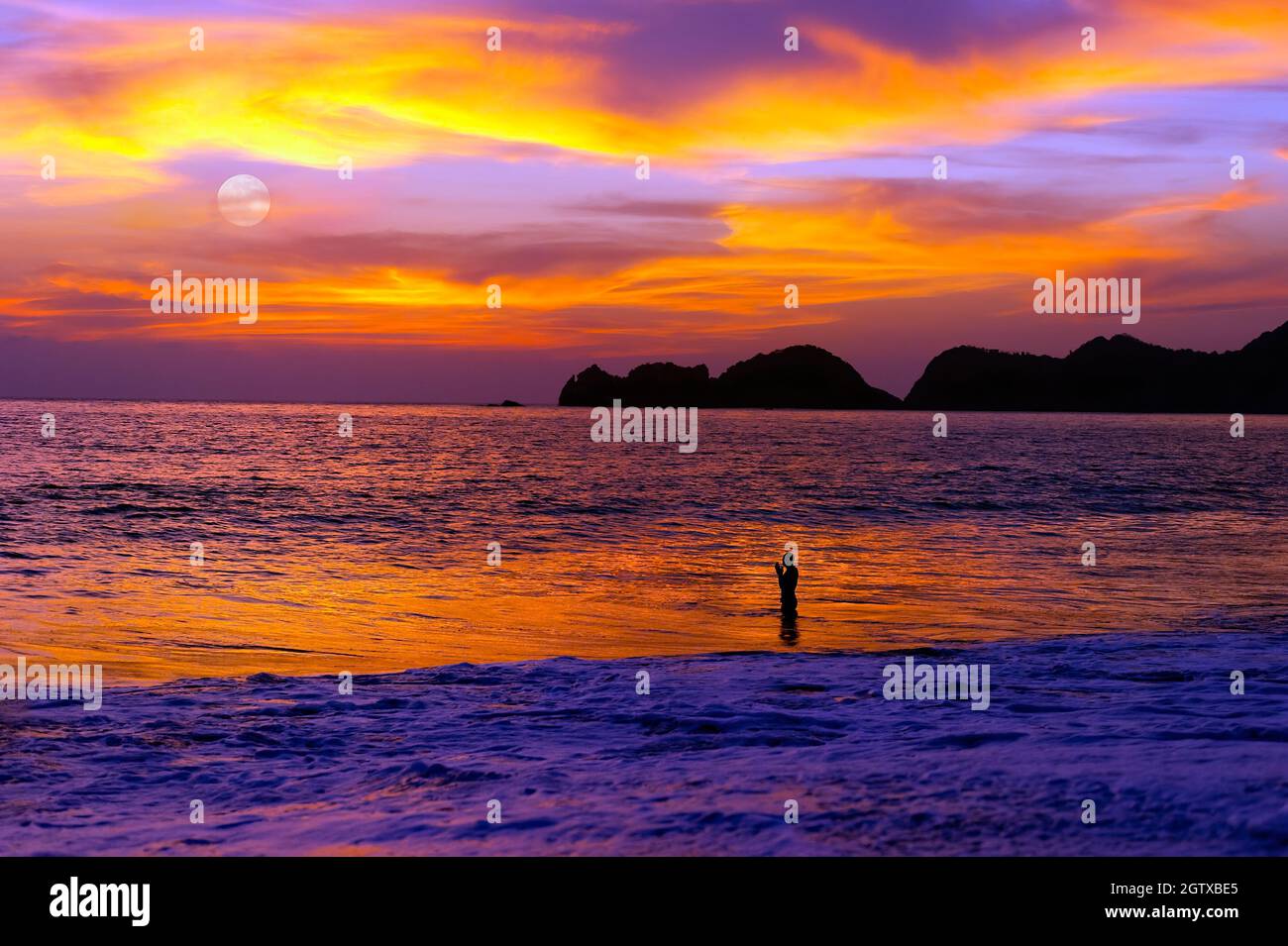 Un homme prie dans l'océan au coucher du soleil alors que la Lune s'élève dans le ciel du soir Banque D'Images