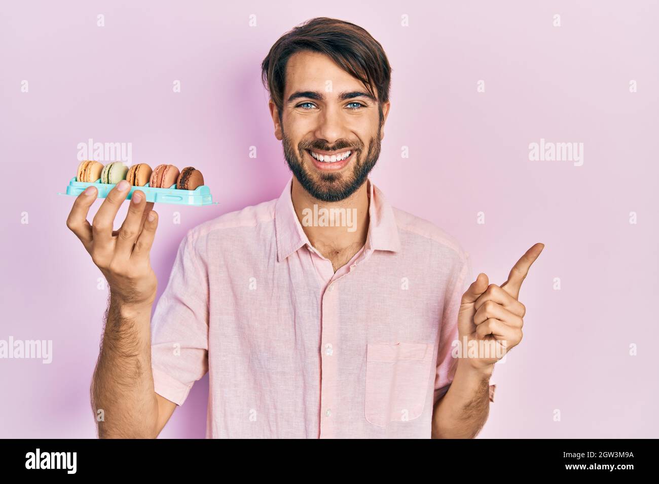 Jeune homme hispanique portant de délicieuses pâtisseries de macrons souriantes pointant avec la main et le doigt sur le côté Banque D'Images