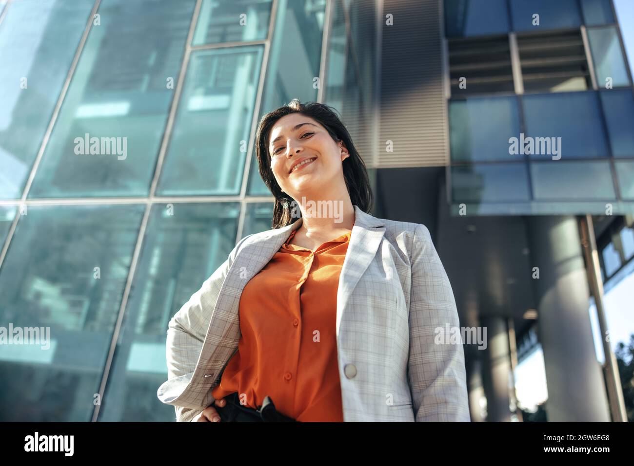 Une femme d'affaires gaie souriant à l'appareil photo tout en se tenant devant un immeuble de bureaux de grande hauteur dans la ville. Femme entrepreneur réussie standi Banque D'Images