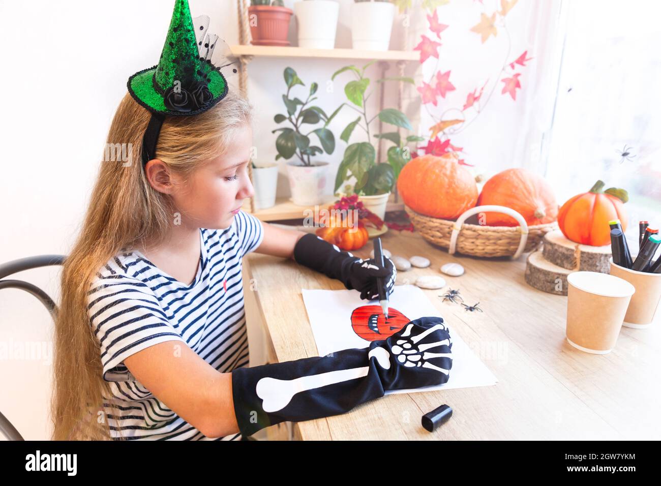Une adolescente dans un chapeau de sorcière vert et des gants noirs avec des os dessine une citrouille, assise près de la fenêtre. Préparation pour la fête d'Halloween. Banque D'Images
