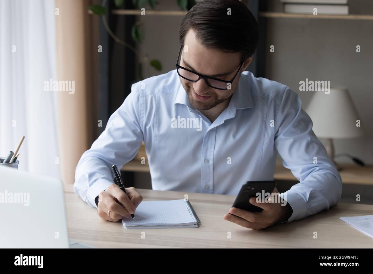 Homme d'affaires souriant dans des lunettes prenant des notes dans un ordinateur portable, tenant un smartphone Banque D'Images