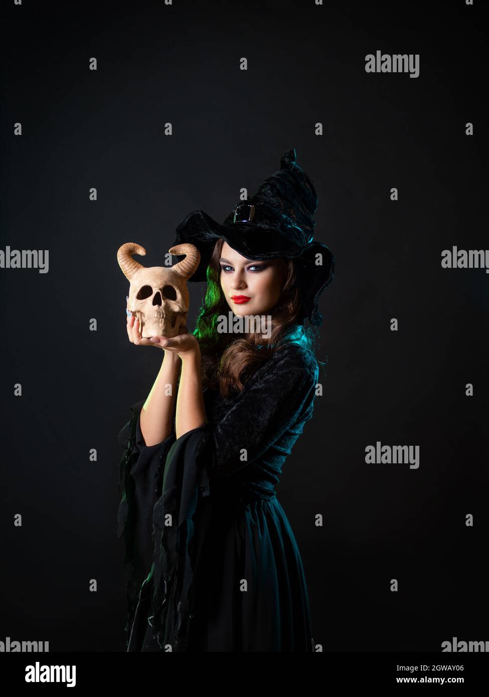 Une belle jeune sorcière tient un crâne de démon corné près de son visage et vous regarde avec un regard malicieux, une invitation à une fête d'Halloween, un costume de sorcière Banque D'Images