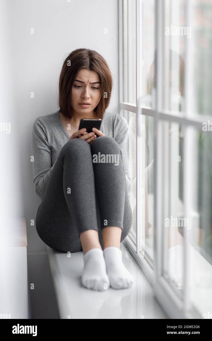 Triste contrarié femme millénaire caucasienne assise sur le rebord de la fenêtre à la maison et lisant un message au téléphone avec de mauvaises nouvelles, souffrant de dépression. Réaction à Banque D'Images