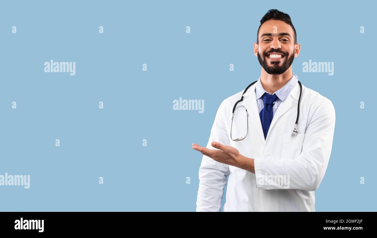Happy Arabic Doctor Man souriant montrant l'espace de copie avec la main Publicité votre texte sur l'arrière-plan Blue Studio. Portrait du médecin ayant réussi. Medi Banque D'Images