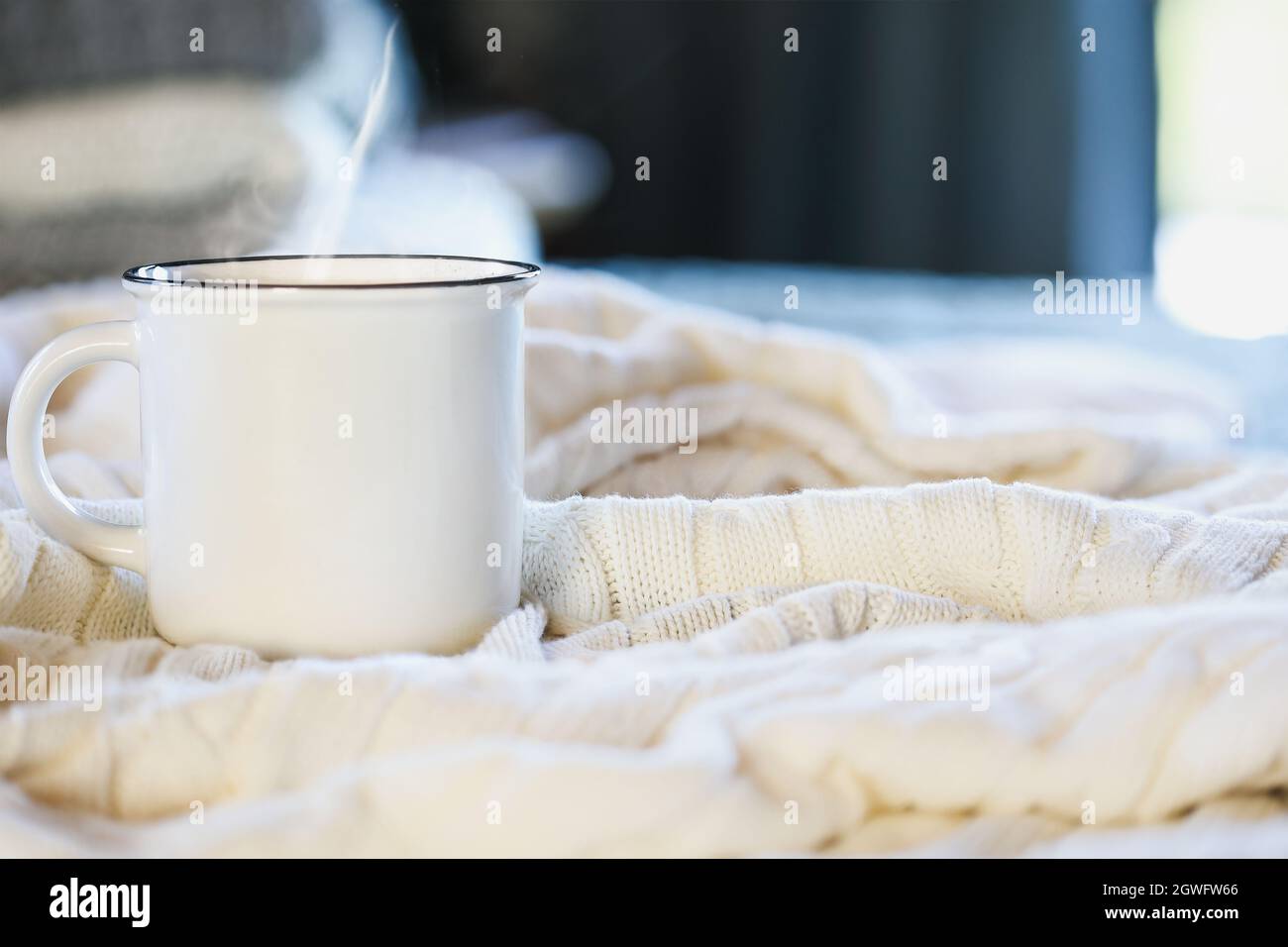 Une tasse de café chaude et chaude, assise sur le dessus d'une couverture en tricot blanc doux sur un lit avec une pile de couvertures en arrière-plan.Mise au point sélective, arrière-plan flou. Banque D'Images