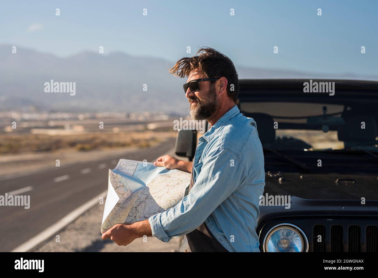 Homme mûr dans des lunettes de soleil en recherchant la direction sur la carte d'emplacement tout en se tenant à côté du capot de la jeep à l'autoroute. Beau homme lisant la carte et regardant awa Banque D'Images